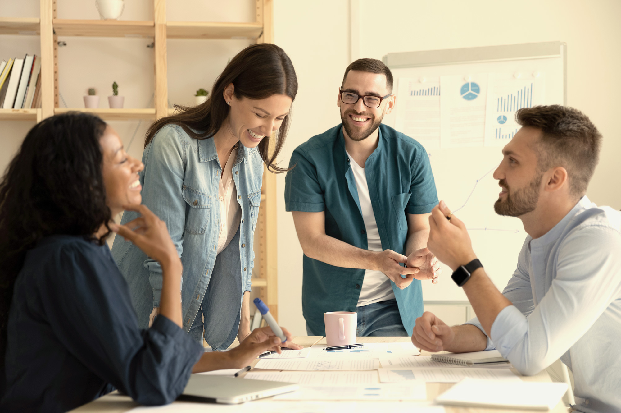  A group of four business professionals are having a cheerful discussion while collaborating on a project in a modern office space.