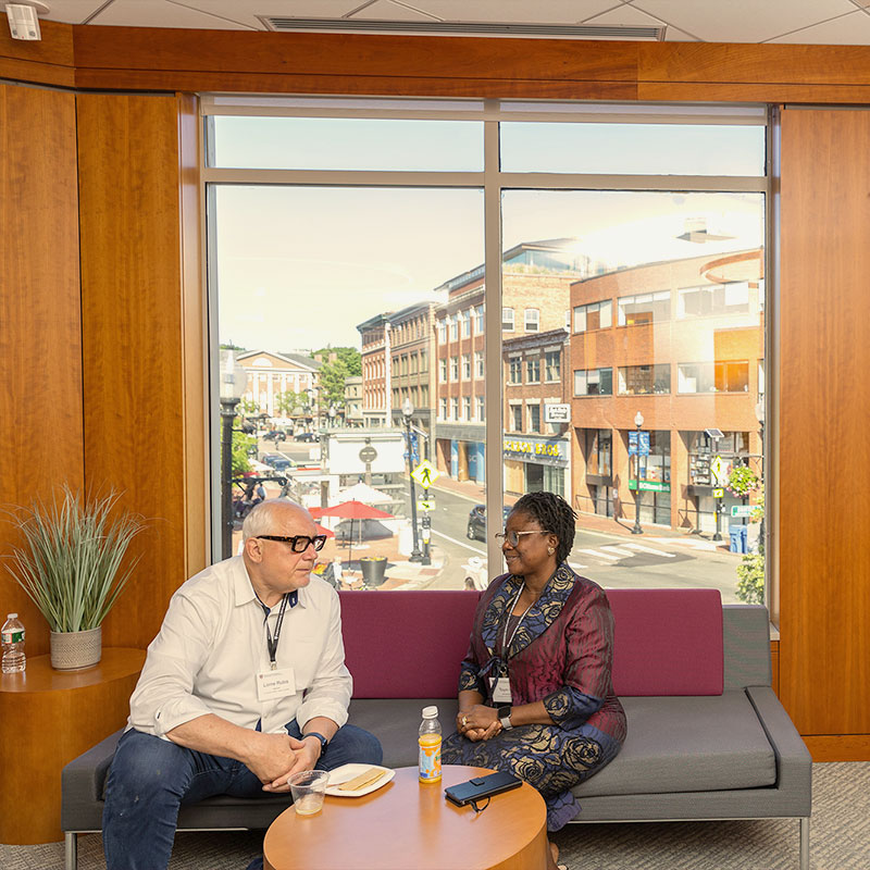 A man and a woman visit on a sofa in front of a window overlooking Harvard Square.